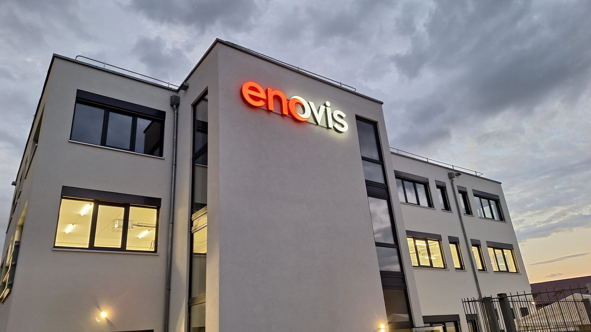 Bild des enovis Standorts Mörsdorf im Sonnenuntergang mit beleuchteten Fenstern und illuminiertem Firmenlogo